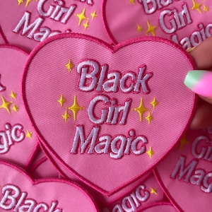 May include: Pink heart-shaped embroidered patches with the text "Black Girl Magic" in white. The patches have a pink border and yellow star accents. The patches are a vibrant and eye-catching accessory.