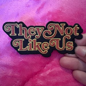 May include: A black and gold embroidered patch with the text "They Not Like Us" in a retro font.