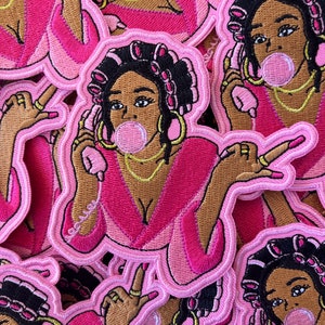 May include: A stack of pink and brown embroidered patches featuring a cartoon woman with pink rollers in her hair, wearing a pink robe and blowing a bubble. The woman is holding a pink phone to her ear.