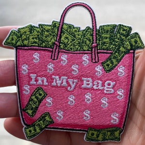 May include: Pink tote bag embroidered with dollar signs and the text "In My Bag". The bag is filled with green cash.
