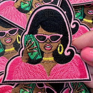 May include: Embroidered iron-on patch featuring a cartoon woman with black hair, pink sunglasses, and a pink outfit. She is holding a stack of green bills to her ear as if on a phone call.