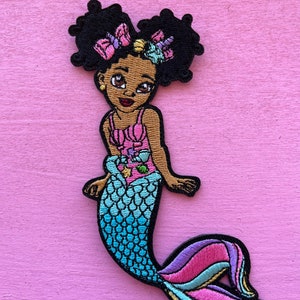 May include: Embroidered patch of a mermaid with dark skin, black curly hair, and a pink top. The mermaid's tail is blue, pink, and purple. The patch has a black border and is set against a pink background.