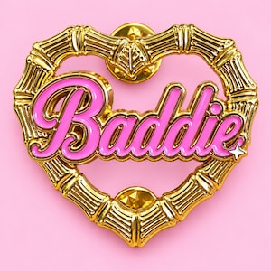 May include: A gold-toned metal pin shaped like a heart with a bamboo-style frame. The center features the word Baddie in bright pink enamel lettering. A small white four-pointed star icon is positioned at the end of the word Baddie.