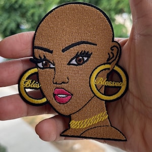 May include: Embroidered patch featuring a stylized Black woman's face with brown skin, brown eyes, and pink lips. The patch includes gold hoop earrings with the word "Blessed" and a gold chain necklace. The patch is outlined in black.