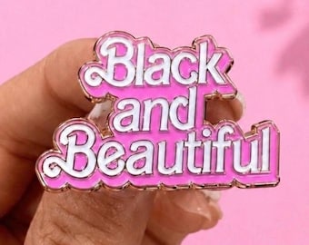 May include: A metal enamel pin featuring the text Black and Beautiful in a white script font with a bright pink background and gold-toned metal edges. The pin is shaped to follow the contours of the lettering, creating a layered, bold appearance.