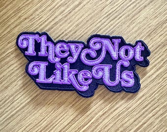 May include: Purple embroidered patch with the text "They Not Like Us" in a retro font style.