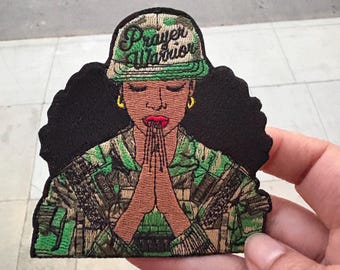 May include: An embroidered patch featuring a woman with dark hair wearing a camouflage jacket and a green baseball cap with the text "Prayer Warrior". She is praying with her hands clasped together.