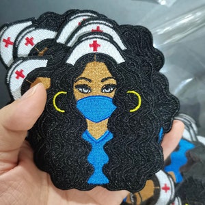 May include: A black and blue embroidered patch featuring a cartoon nurse with curly black hair, wearing a blue surgical mask, a white nurse's cap with a red cross, and gold hoop earrings.