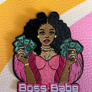 May include: Embroidered iron-on patch featuring a cartoon woman with black hair holding stacks of money in each hand. She is wearing a pink dress and the text "Boss Babe" is below her.
