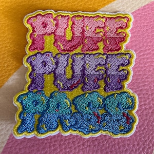 May include: A colorful embroidered patch with the words "Pure Puff" in a bubble-like font. The letters are outlined in yellow and have a dripping effect. The patch is on a pink and yellow striped background.