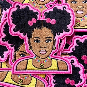 May include: Embroidered patch featuring a cartoon girl with brown skin, black curly hair, and pink bows. She wears a yellow top and a colorful beaded necklace. The patch has a pink outline.