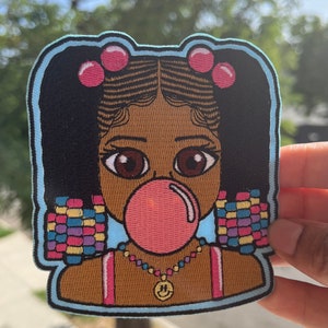 May include: Embroidered patch featuring a cartoon girl with brown skin, black hair in pigtails with pink bows, wearing a pink tank top, a beaded necklace with a smiley face charm, and blowing a pink bubble. The girl is surrounded by a blue border and colorful beads.