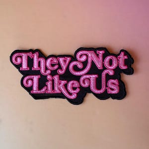 May include: A black embroidered patch with the phrase "They Not Like Us" in pink, stylized lettering. The patch has a black border and is likely designed to be sewn or ironed onto fabric.