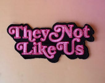 May include: A black embroidered patch with the phrase "They Not Like Us" in pink, stylized lettering. The patch has a black border and is likely designed to be sewn or ironed onto fabric.