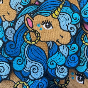 May include: A close-up of a stack of embroidered patches featuring a unicorn with blue and light blue hair, brown fur, and a gold horn. The unicorn has a pink ear and a large, brown eye with blue glitter.