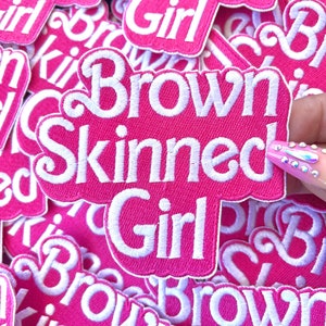 May include: A close-up of several pink fabric patches with white embroidered text that reads "Brown Skinned Girl".