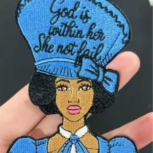 May include: Embroidered patch featuring a woman in a blue hat and dress. The patch includes the text "God is within her She not fail" in cursive script. The woman has dark skin, black hair, and pink lips.