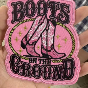 May include: Pink embroidered patch with the words "BOOTS ON THE GROUND" in black script. The patch features a pink oval with two pink and white cowboy boots. Gold stars and a gold rope border the oval.