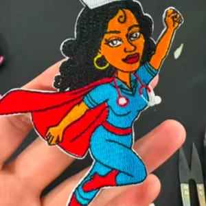 May include: Embroidered patch of a superhero nurse with a red cape, blue scrubs, and a white nurse's cap with a red cross. The nurse has dark hair, brown skin, and a stethoscope around her neck.