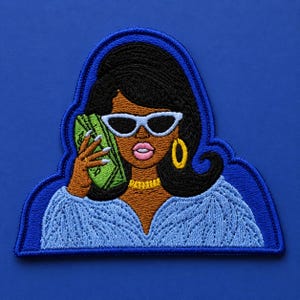 May include: Embroidered patch featuring a Black woman with sunglasses holding a wad of cash to her ear. She wears a blue sweater, gold jewelry, and has black hair. The patch is outlined in blue.