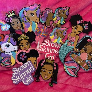 May include: A collection of colorful embroidered patches featuring Black girls, unicorns, mermaids, and the text "Brown Skinned Girl." Patches include rainbows, lollipops, and crowns. The patches are on a pink background.