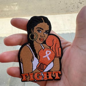 May include: Orange and white embroidered patch of a woman wearing boxing gloves with a white ribbon on one glove. The text "FIGHT" is embroidered below the woman.