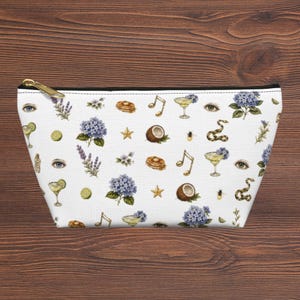 Puede incluir: Un estuche blanco con cremallera que presenta un patrón repetitivo de hortensias azules, ramas de lavanda, rodajas de lima, cócteles, notas musicales, ojos humanos, serpientes, abejas, mitades de coco, estrellas y pilas de tortitas.
