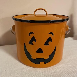 Può includere: Secchio di Halloween in metallo arancione con coperchio e manico. Il secchio presenta una faccia di jack-o'-lantern con occhi, naso e bocca sorridente neri. Il bordo e il manico sono neri.
