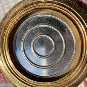 May include: Open, gold-toned compact with a silver interior. The interior features a series of concentric circles. The compact is open, revealing the mirror and powder well. The patent number 764125 is visible on the hinge.