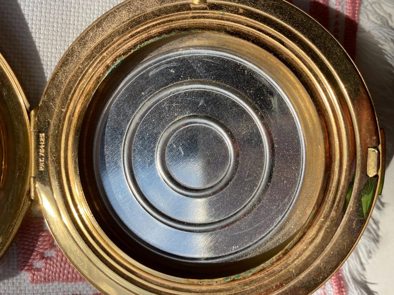 May include: Open, gold-toned compact with a silver interior. The interior features a series of concentric circles. The compact is open, revealing the mirror and powder well. The patent number 764125 is visible on the hinge.