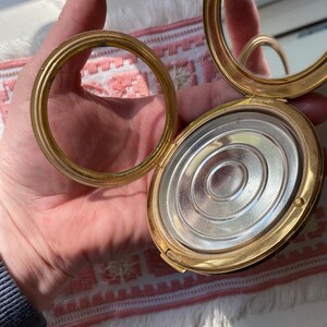 May include: Open, gold-toned compact with a mirror and a separate ring. The compact's interior features a silver-colored, circular design. The compact is held open, revealing the mirror and the powder compartment. The ring is detached.