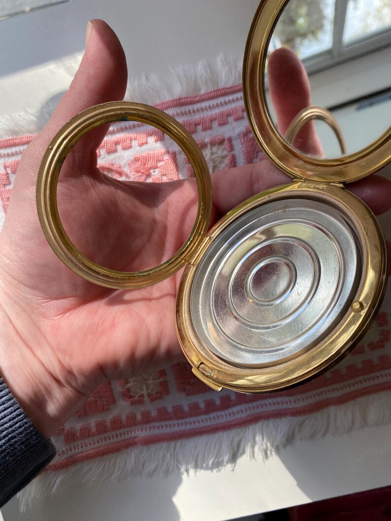 May include: Open, gold-toned compact with a mirror and a separate ring. The compact's interior features a silver-colored, circular design. The compact is held open, revealing the mirror and the powder compartment. The ring is detached.