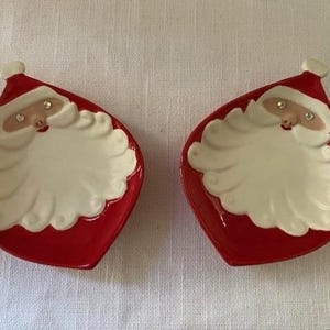 May include: Two red, teardrop-shaped dishes featuring Santa Claus faces. Each dish has a white beard, a rosy face, and a red hat with a white pom-pom. The dishes are likely for serving small snacks or candies during the holiday season.