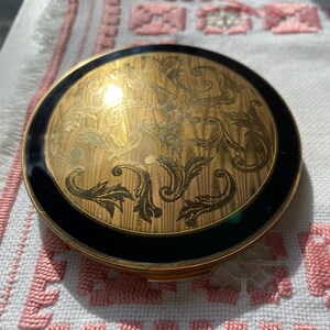 May include: A round, vintage compact with a gold-tone finish and intricate engraved floral design. The compact features a black enamel border. The compact is a classic beauty accessory.