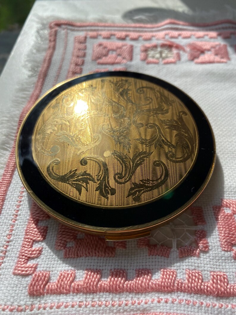 May include: A round, vintage compact with a gold-tone finish and intricate engraved floral design. The compact features a black enamel border. The compact is a classic beauty accessory.