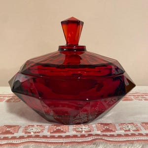 May include: A deep red, faceted glass candy dish with a matching lid. The bowl features a geometric, multi-sided design with sharp angles and a pointed finial on top of the lid. The glass is translucent, allowing light to pass through the red color.