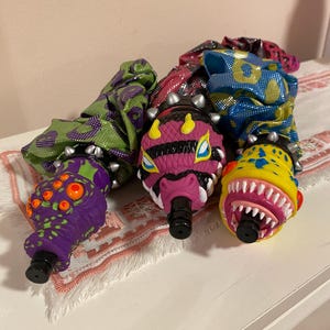 Puede incluir: Tres tapones de botella con temática de monstruos. Los tapones presentan un monstruo morado con múltiples ojos, una cabeza rosa con forma de serpiente y un monstruo amarillo con dientes afilados. Cada tapón tiene una base negra y un collar con tachuelas.