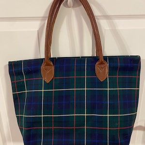 May include: A tote bag with a green, blue, red, and yellow plaid pattern. The bag has brown leather handles and accents. The bag is rectangular and appears to be made of a sturdy fabric.