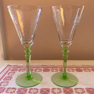 May include: Two clear glass wine glasses with etched floral designs. The stems and bases are a vibrant green. The glasses are displayed on a white and pink embroidered placemat.
