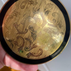 May include: A vintage gold compact with a black border. The compact features an ornate, etched floral design on a textured background. The compact is round and appears to be made of metal.