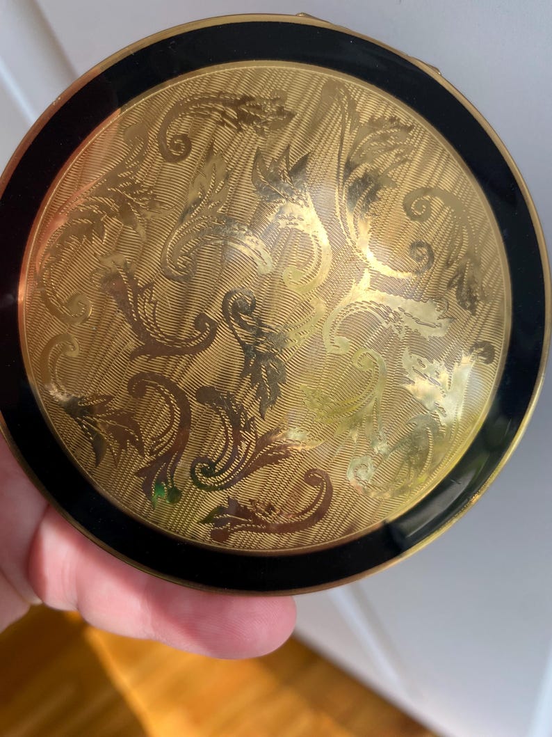 May include: A vintage gold compact with a black border. The compact features an ornate, etched floral design on a textured background. The compact is round and appears to be made of metal.
