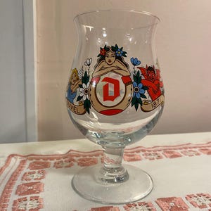 May include: A clear glass beer glass with a wide bowl and a stem. The glass is decorated with colourful illustrations of a woman, an angel, and a devil, and the text "DUVEL 1871". It sits on a white and pink embroidered cloth.