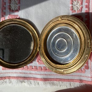 May include: Open gold-toned compact with a mirror on one side and a silver-toned powder compartment on the other. The compact is round and has a hinged closure. The mirror is circular.