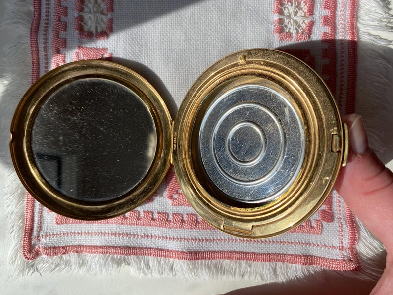 May include: Open gold-toned compact with a mirror on one side and a silver-toned powder compartment on the other. The compact is round and has a hinged closure. The mirror is circular.