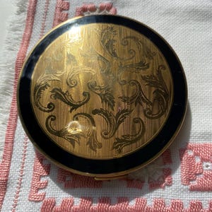 May include: A round, vintage compact with a gold-toned, ornate floral design on the lid. The compact has a black border and is set against a white and pink embroidered fabric.