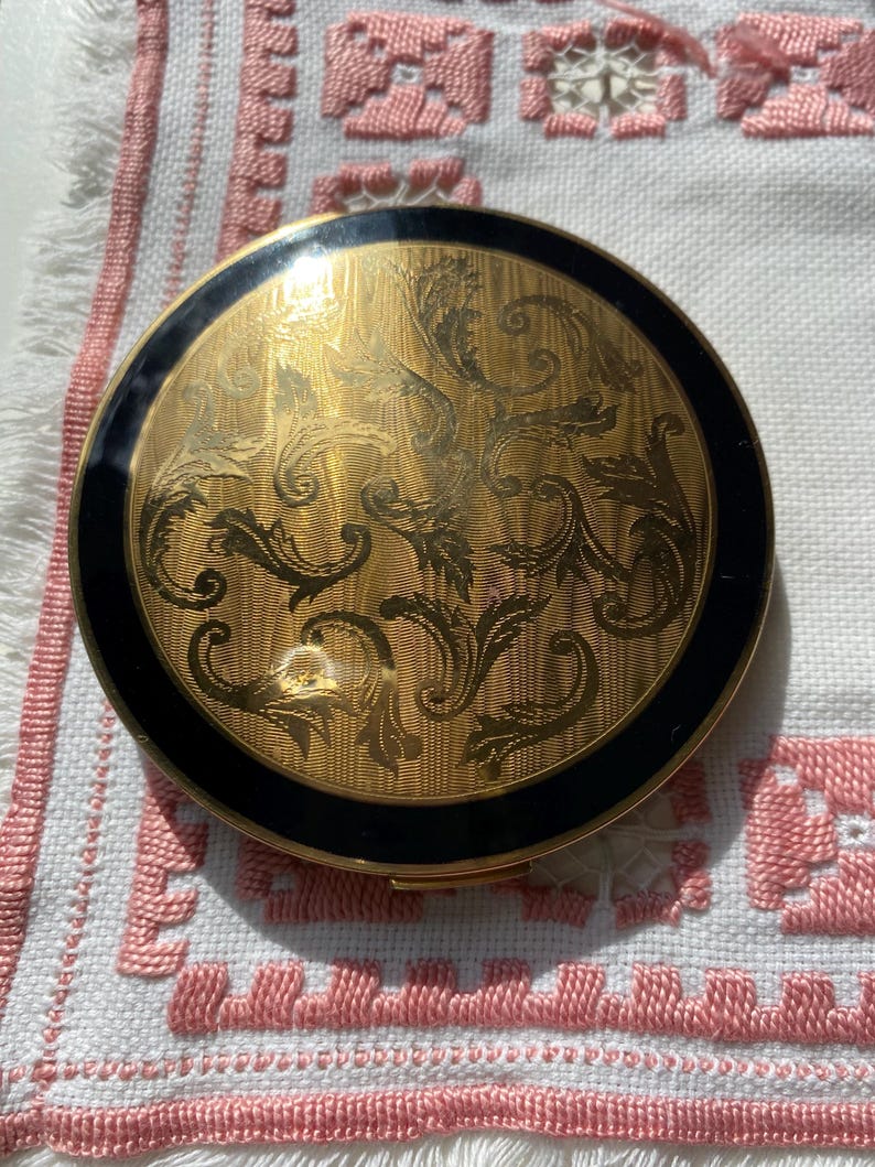 May include: A round, vintage compact with a gold-toned, ornate floral design on the lid. The compact has a black border and is set against a white and pink embroidered fabric.