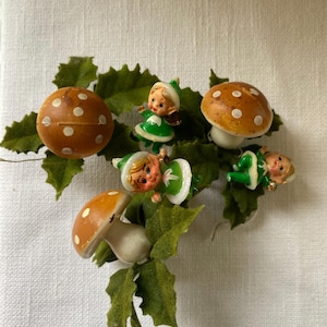 Puede incluir: Una colección de adornos navideños vintage con figuras en miniatura de setas y elfos. Las setas tienen sombreros marrones con lunares blancos. Los elfos visten trajes verdes y blancos. Los adornos están dispuestos con acentos de hojas verdes.