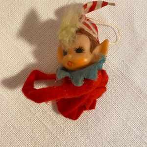 Puede incluir: Un adorno de elfo navideño vintage. El elfo tiene una cara pintada, cabello rubio y un sombrero a rayas rojas y blancas. Lleva un cuello azul y un atuendo de fieltro rojo. Se adjunta una cuerda dorada para colgar.