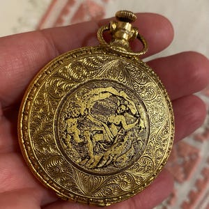 May include: A gold-colored pocket watch with intricate floral and figure engravings. The watch features a detailed design on the front and a winding stem at the top. The watch is round and appears to be vintage.