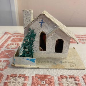 Puede incluir: Una pequeña maqueta de iglesia de papel blanco con una cruz azul en el tejado y la inscripción "Sunday School" en la base. La iglesia tiene dos ventanas arqueadas y una chimenea. Un pequeño árbol verde está en el lateral.